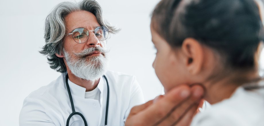 Little girl have a visit. Senior otorhinolaryngologist with grey hair and beard in white coat is indoors in clinic